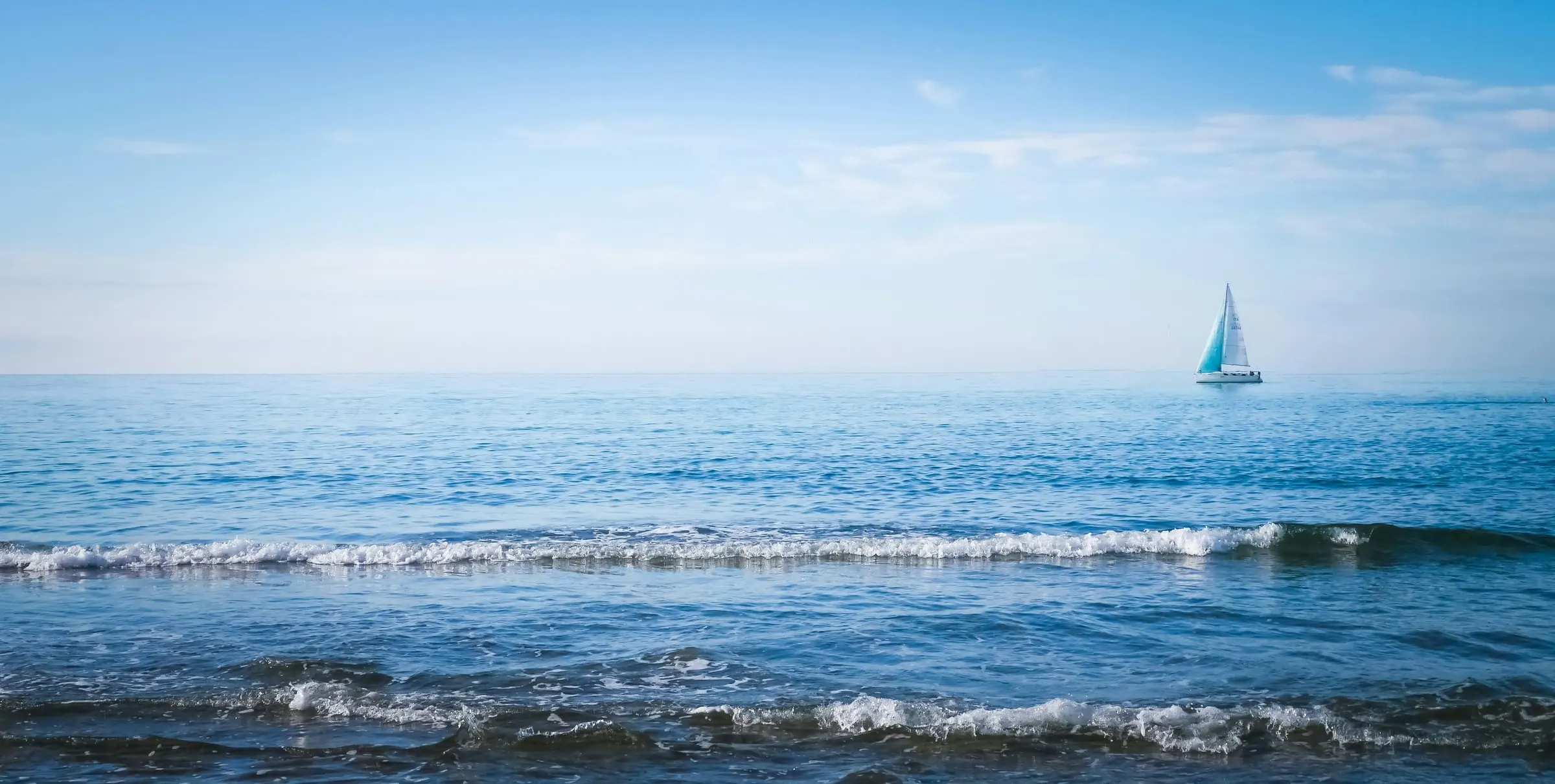 Calm open sea under a wide blue sky with a small teal-sailed boat on the horizon