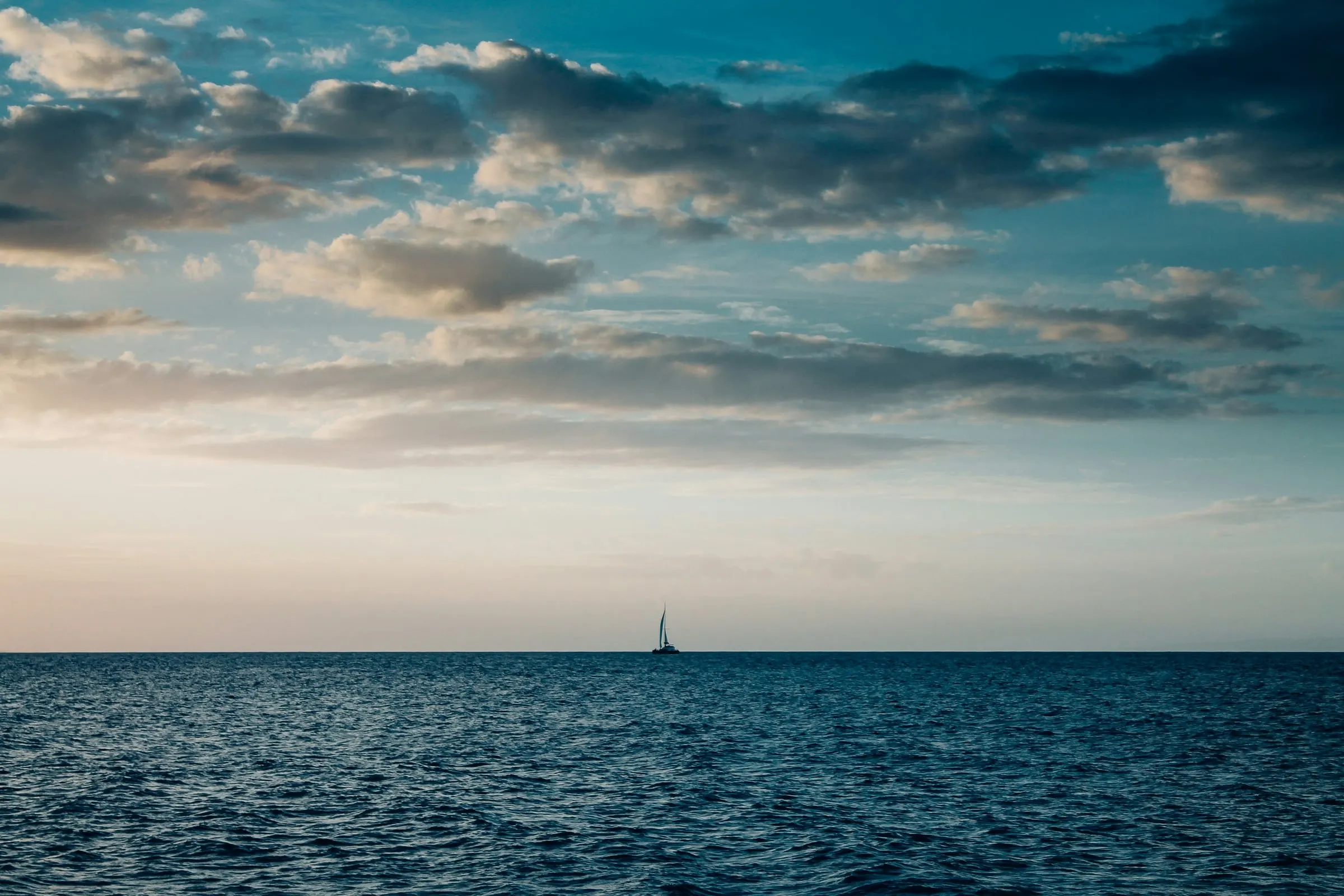 Open ocean with a small sailboat on the horizon beneath scattered clouds